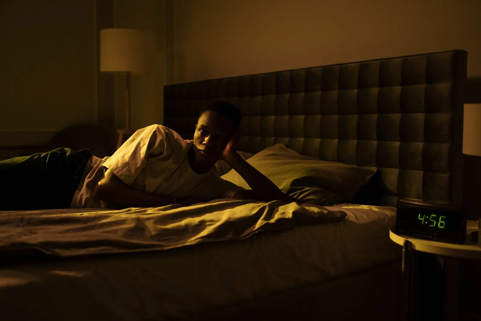 A man lying awake in bed in the early morning beside a clock showing 4:56 a.m.