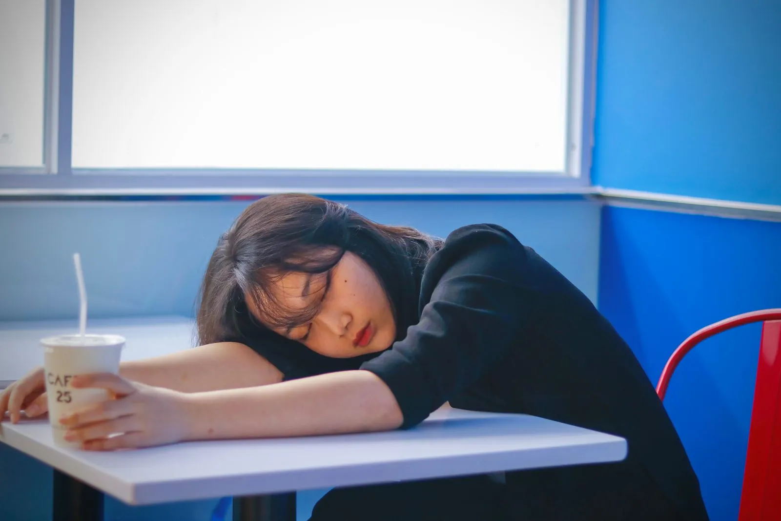A tired woman resting her head on a café table, holding a takeaway coffee cup