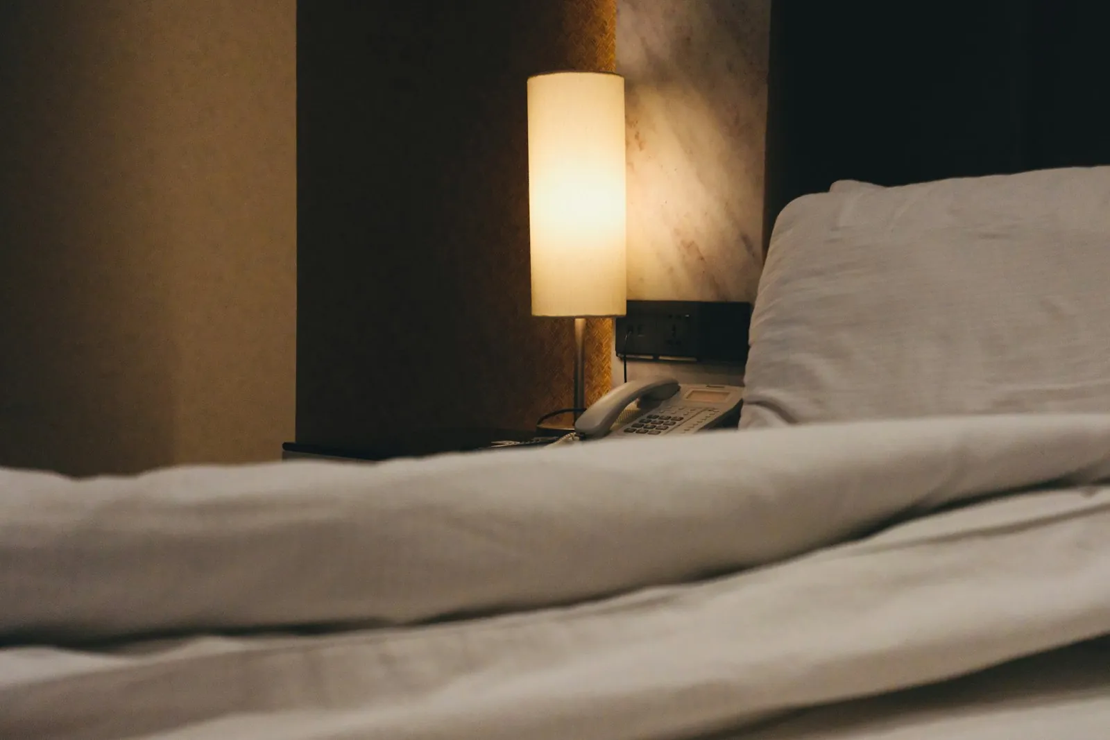 A bedside lamp glowing beside a rumpled white bed at night