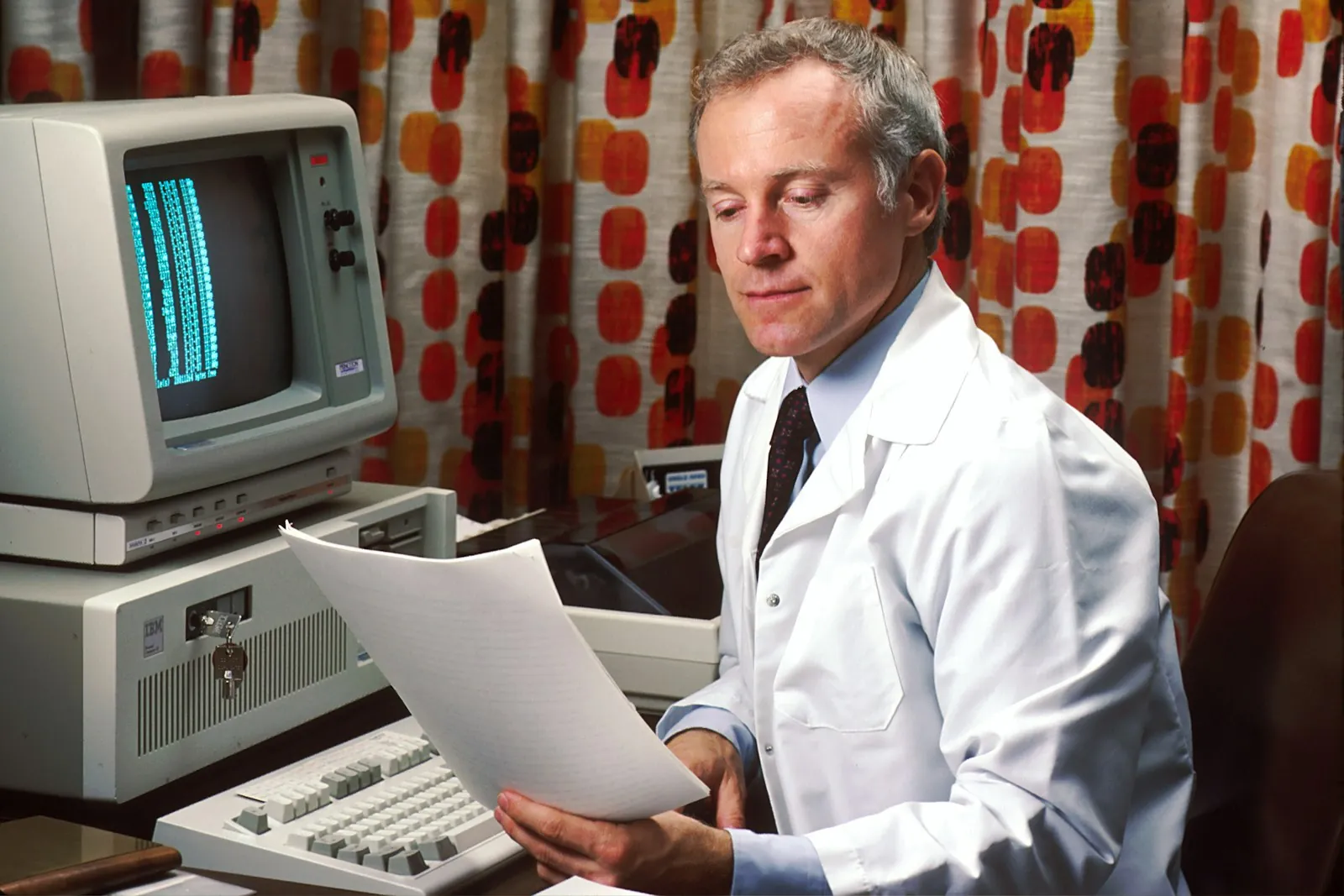 A man reads printed papers at his desk beside a computer in 1987