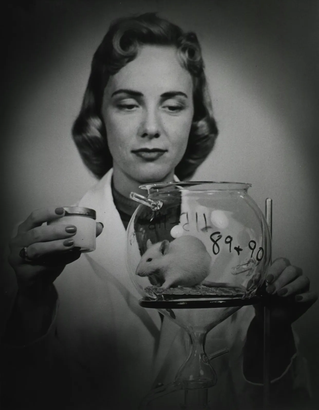 Black-and-white photo of a woman scientist in a lab coat looking at a white mouse inside a labeled glass jar