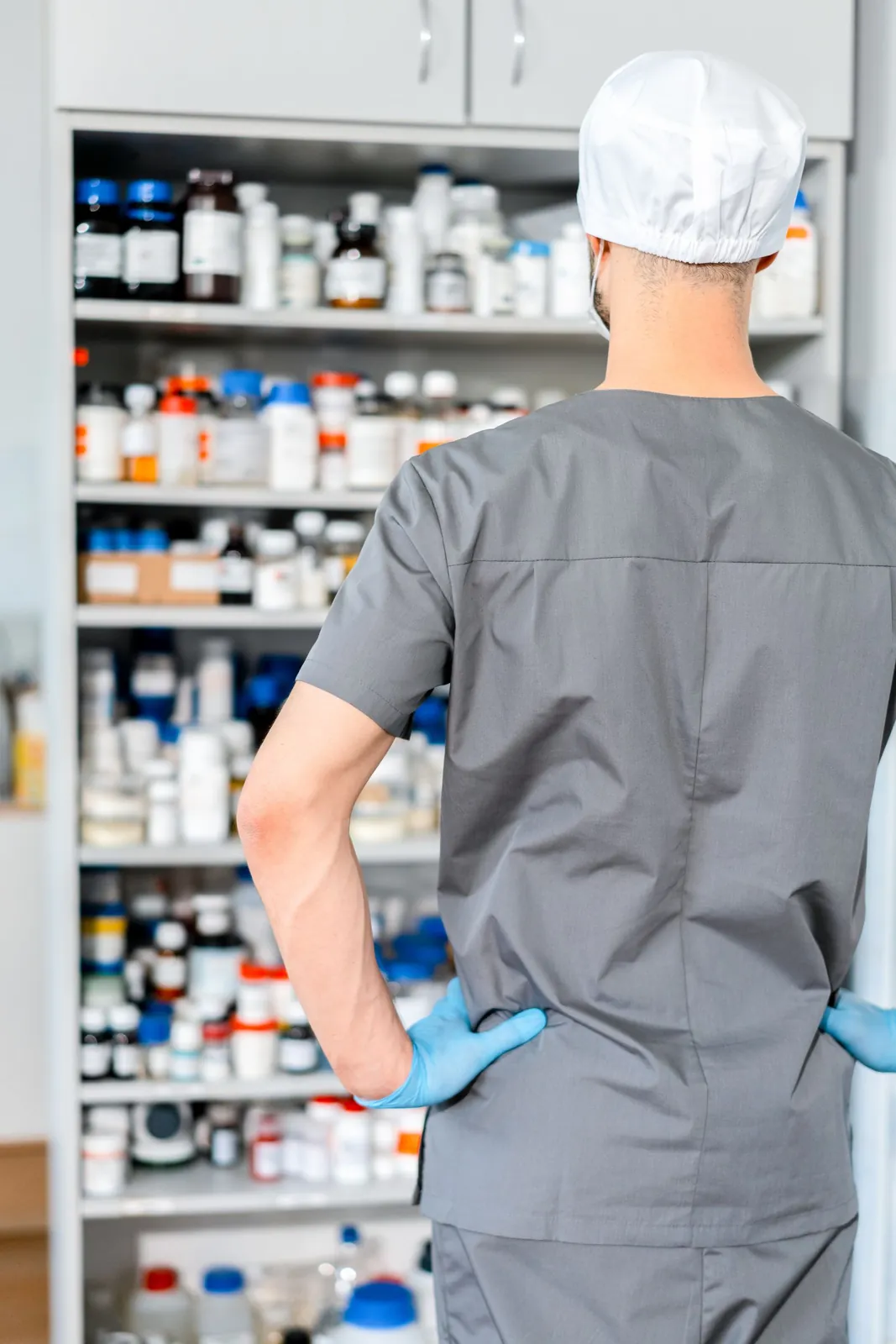 A pharmacist in gloves, seen from behind, sorting medicine on pharmacy shelves